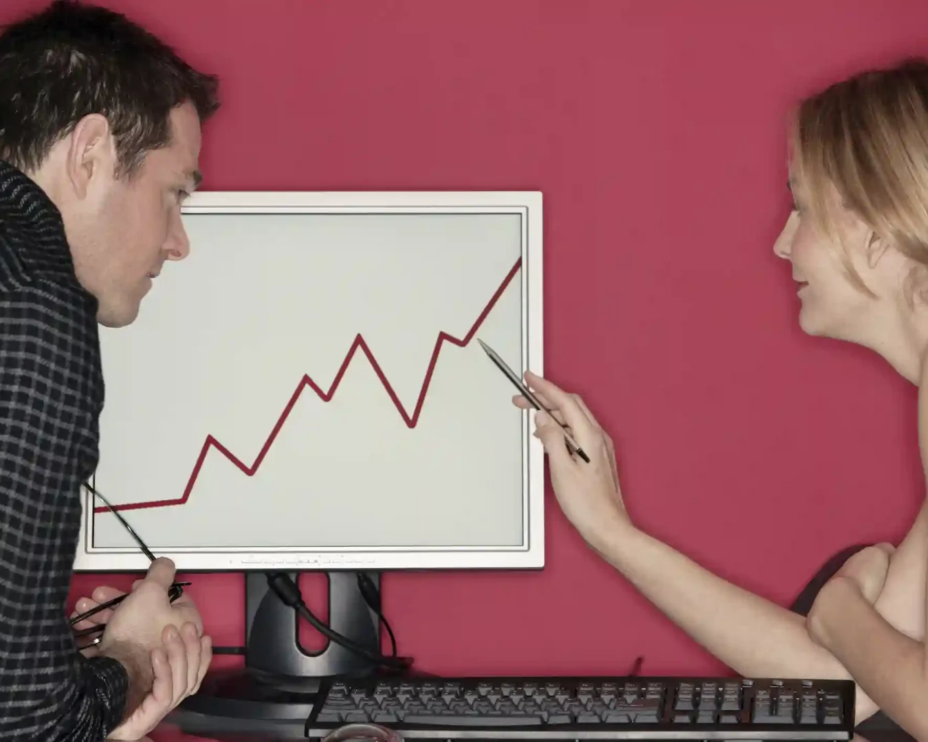 Ein Mann und eine Frau diskutieren über ein Liniendiagramm, das einen Aufwärtstrend auf einem Computermonitor zeigt - vielleicht um ihre justtrade erfahrung zu überprüfen. Die Frau zeigt mit einem Stift auf den Bildschirm, während der Mann zuhört und seine Brille festhält. Der Hintergrund ist einfarbig rosa-rot.