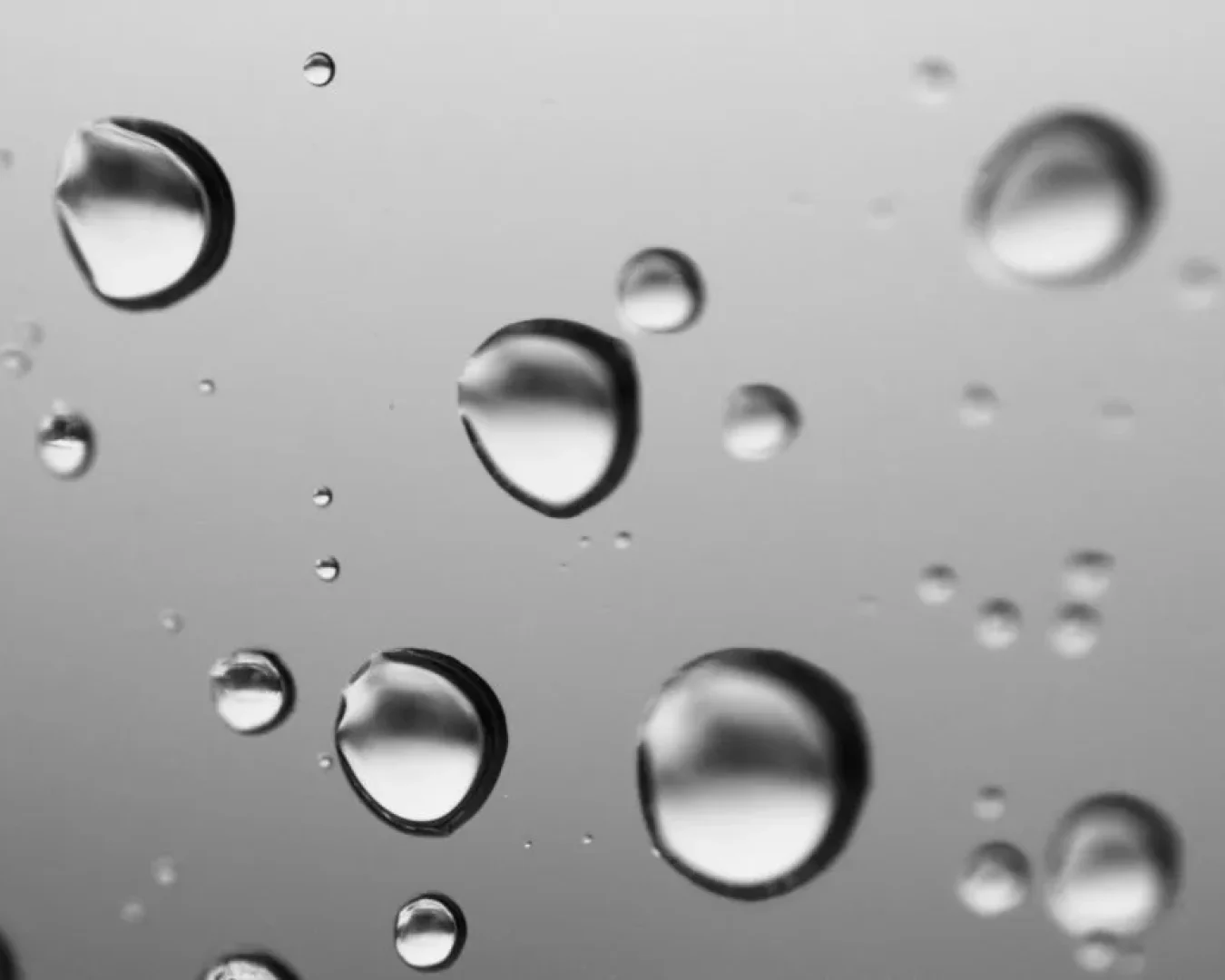 Nahaufnahme von Wassertröpfchen auf einer Glasoberfläche aus AISI 316L vor einem unscharfen grauen Hintergrund. Die Tröpfchen variieren in ihrer Größe und reflektieren das Licht, wodurch ein weiches, abstraktes Muster entsteht.