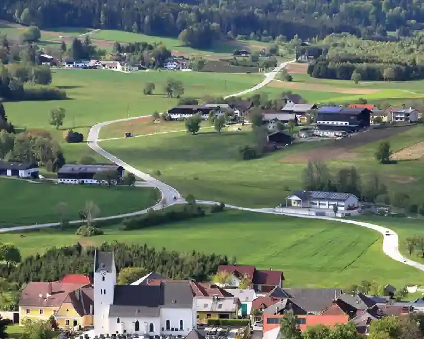 Eine kurvenreiche Landstraße schlängelt sich durch eine üppig grüne Landschaft mit verstreuten Häusern, Bauernhöfen, Feldern und Bäumen, alles vor dem Hintergrund bewaldeter Hügel - und spiegelt den Geist der Energiewende in dem friedlichen Dorf mit seiner malerischen Kirche im Vordergrund wider.