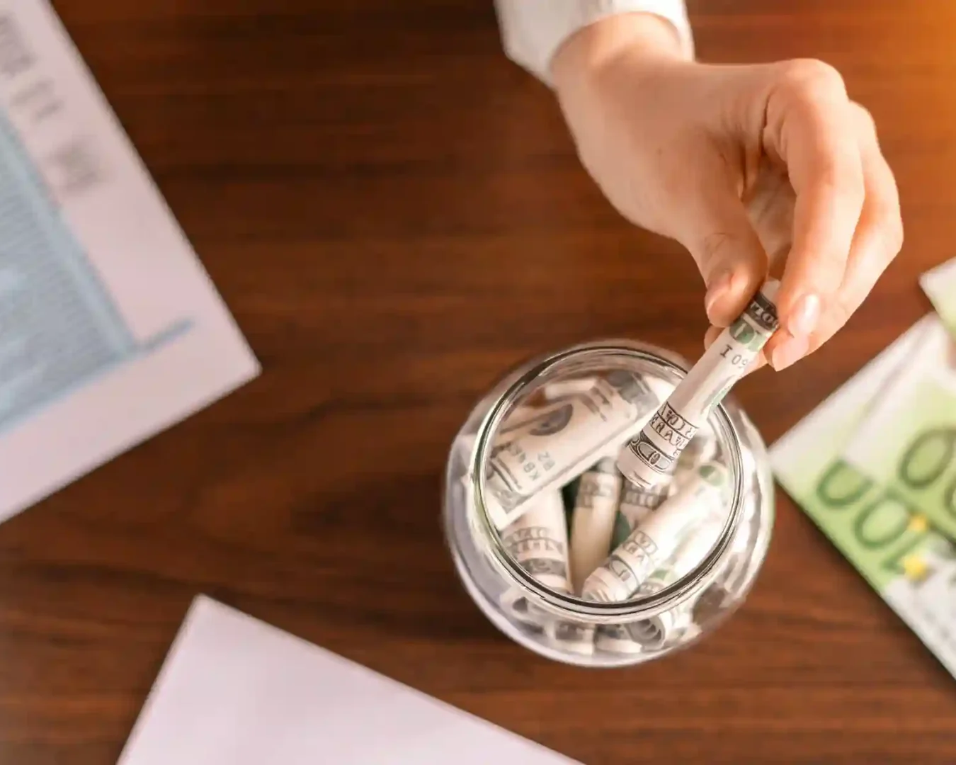 Die Hand einer Person legt einen gerollten US-Dollar-Schein in ein Glasgefäß, das mit weiteren gerollten Scheinen, Dokumenten, Euro-Banknoten und einem Tagesgeld-ETF-Auszug auf einem Holztisch gefüllt ist.