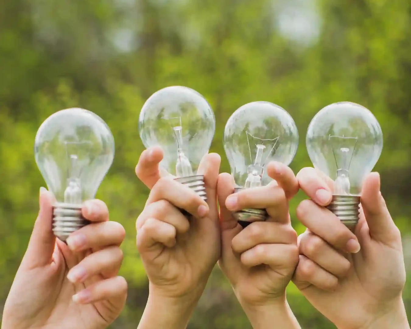 Vier Hände halten klare Glühbirnen vor einem unscharfen grünen Hintergrund, der Ideen, Kreativität oder nachhaltige Energie symbolisiert - ähnlich wie bei der Abwägung der Vorteile von Tagesgeld und Festgeld bei der Wahl von Finanzlösungen.