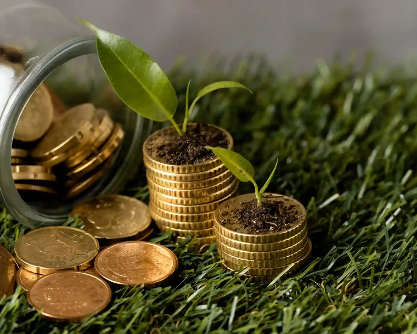 Ein Glasgefäß verschüttet Goldmünzen auf Gras; zwei Münzstapel, auf denen Erde und kleine Grünpflanzen wachsen, symbolisieren nachhaltiges Tagesgeld und verdeutlichen finanzielles Wachstum durch nachhaltige Investitionen.