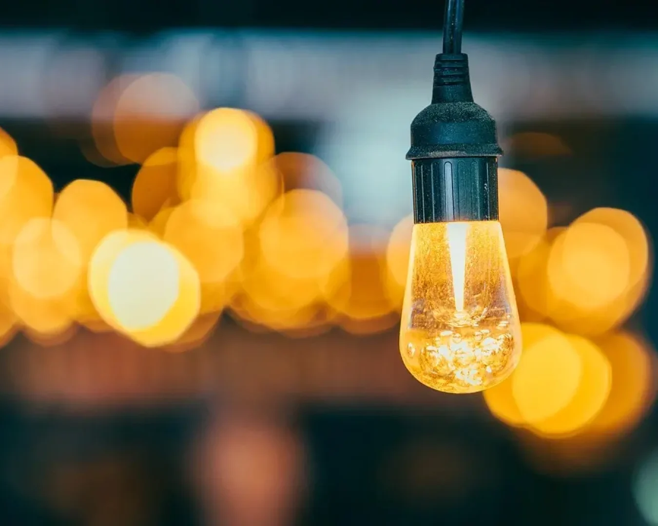 Eine Nahaufnahme einer hängenden Glühbirne, die warm leuchtet, mit vielen unscharfen gelben und orangefarbenen Lichtern im Hintergrund, die einen Bokeh-Effekt erzeugen - perfekt für die Veranschaulichung von Energie- oder Stromvergleichskonzepten in geschäftlichen Zusammenhängen.