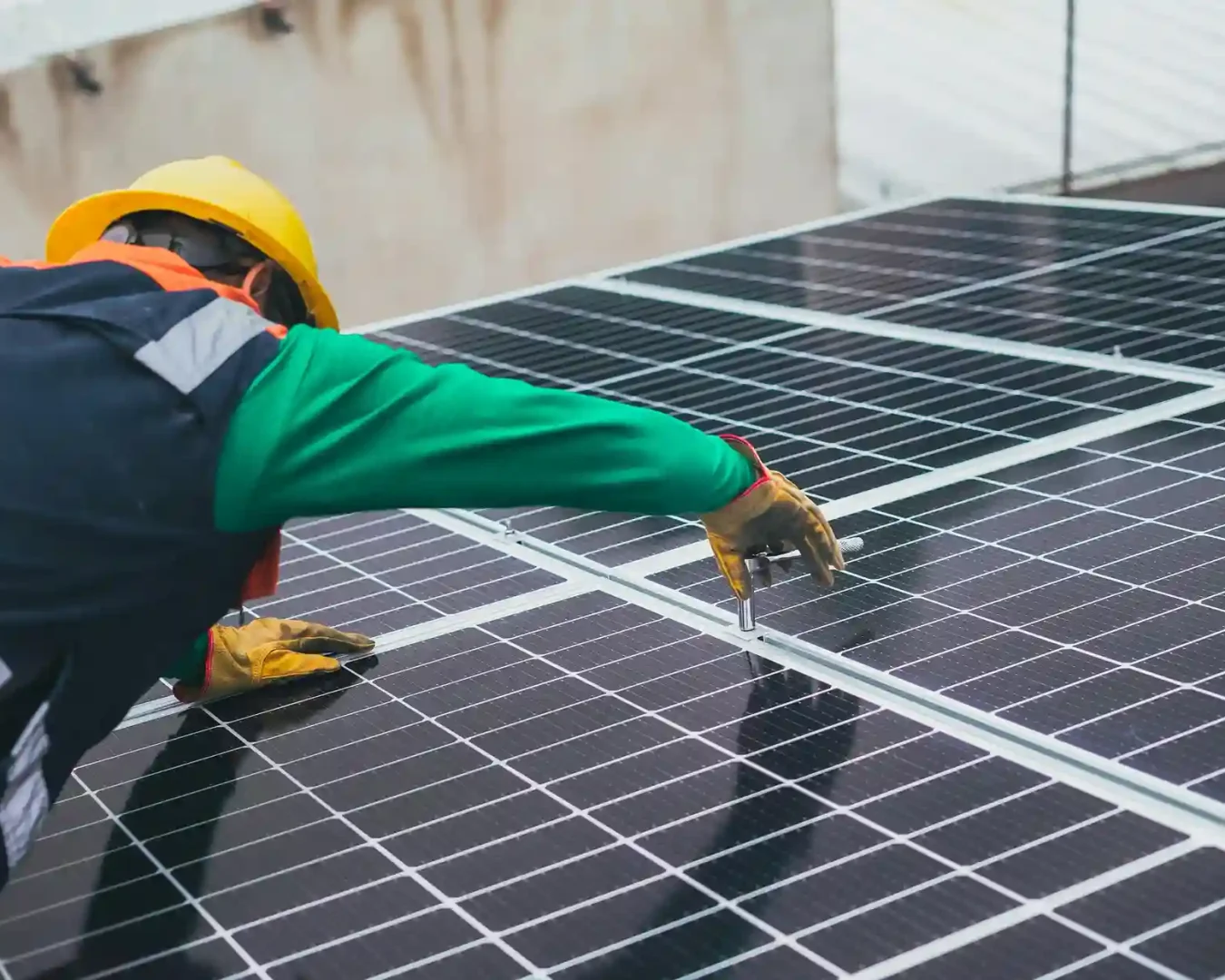 Ein Arbeiter, der einen gelben Schutzhelm und eine Sicherheitsweste trägt, installiert oder wartet Solarmodule auf einem Dach und benutzt ein Werkzeug, um die Befestigungen der Module zu justieren - ein Beispiel dafür, wie eine Energiegenossenschaft nachhaltige Energielösungen unterstützen kann.