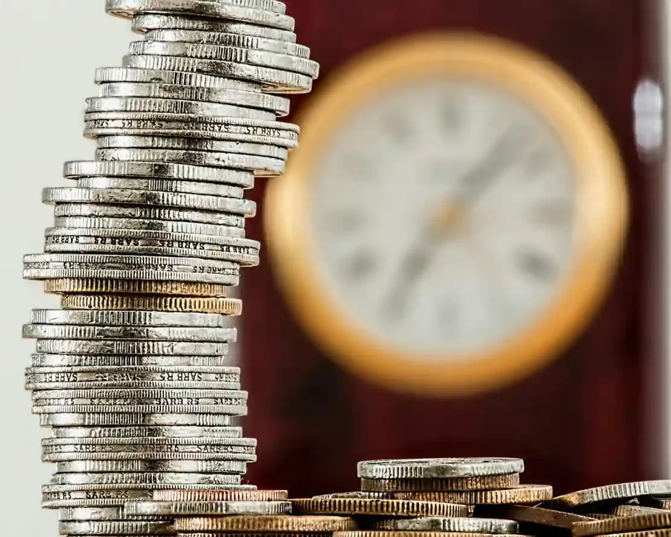 Im Vordergrund ist ein hoher, ungleichmäßiger Stapel Silbermünzen zu sehen, im Hintergrund eine unscharfe Uhr, die das Konzept von Zeit und Geld veranschaulicht - ideal für die Diskussion von Tagesgeld- und Festgeldoptionen.