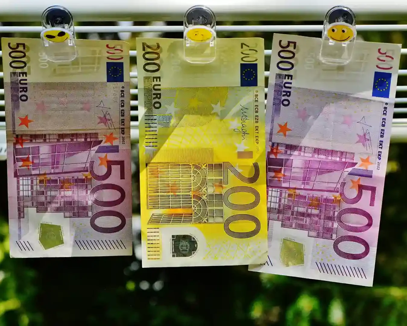 Drei Euro-Banknoten (zwei 500er und ein 200er) hängen auf einer Linie mit Smiley-Klammern, die an Geldwäsche erinnern. Der unscharfe grüne Hintergrund deutet die Natur an - ein Bild, das so kontrastreich ist wie die Wahl zwischen Tagesgeld und Festgeld für Ihre Ersparnisse.