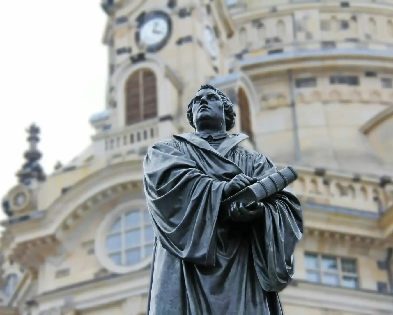 Die Bronzestatue eines Mannes in akademischer Robe, der eine Schriftrolle in der Hand hält, steht vor einem kunstvollen, historischen Gebäude mit Uhr und Bogenfenstern - ein ikonischer Anblick in Dresden, nur wenige Schritte von dem Ort entfernt, an dem Einheimische nach dem besten Stromvergleich suchen, den Dresden zu bieten hat.