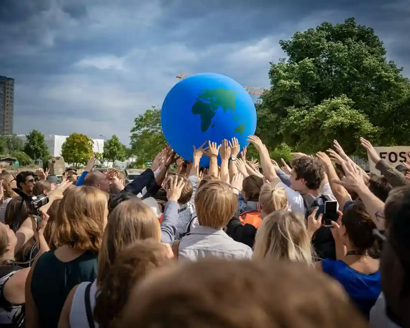 Eine große Gruppe von Menschen erhebt im Freien ihre Hände, um einen riesigen aufblasbaren Globus zu stützen, der die Einheit symbolisiert - ähnlich wie die Wahl zwischen Tagesgeld und Festgeld für kollektive finanzielle Ziele - mit Bäumen und Gebäuden im Hintergrund unter einem bewölkten Himmel.