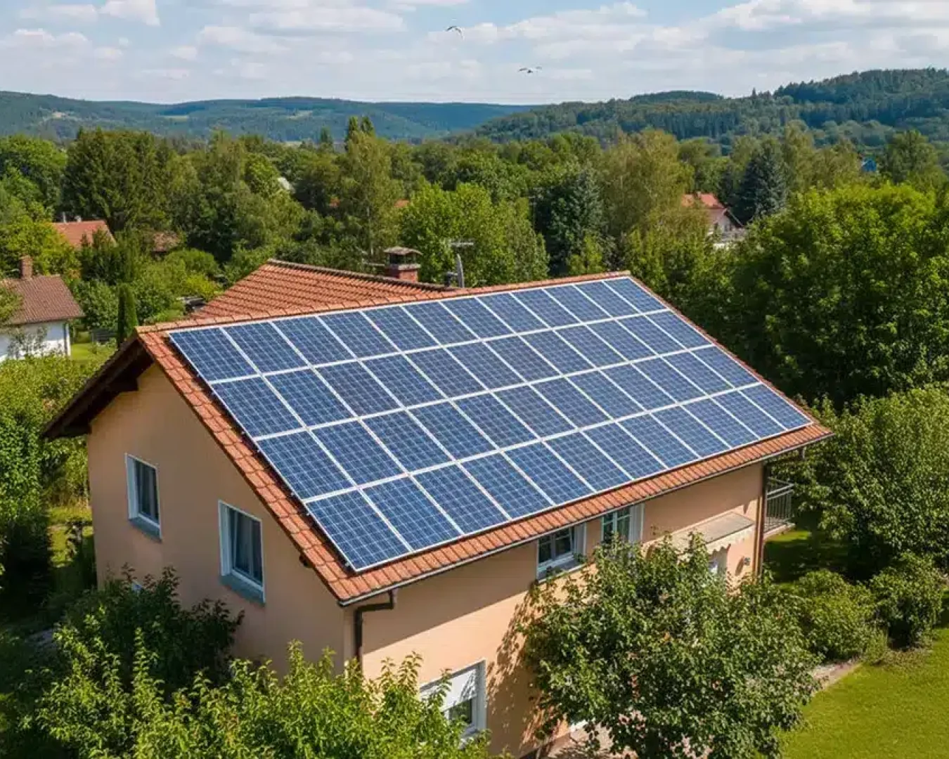 Ein Haus mit einem roten Ziegeldach, das mit Solarzellen bedeckt ist, ist von Bäumen und Grünflächen in einer ländlichen Gegend umgeben. Im Hintergrund sind ein Kirchturm und bewaldete Hügel unter einem teilweise bewölkten Himmel zu sehen.