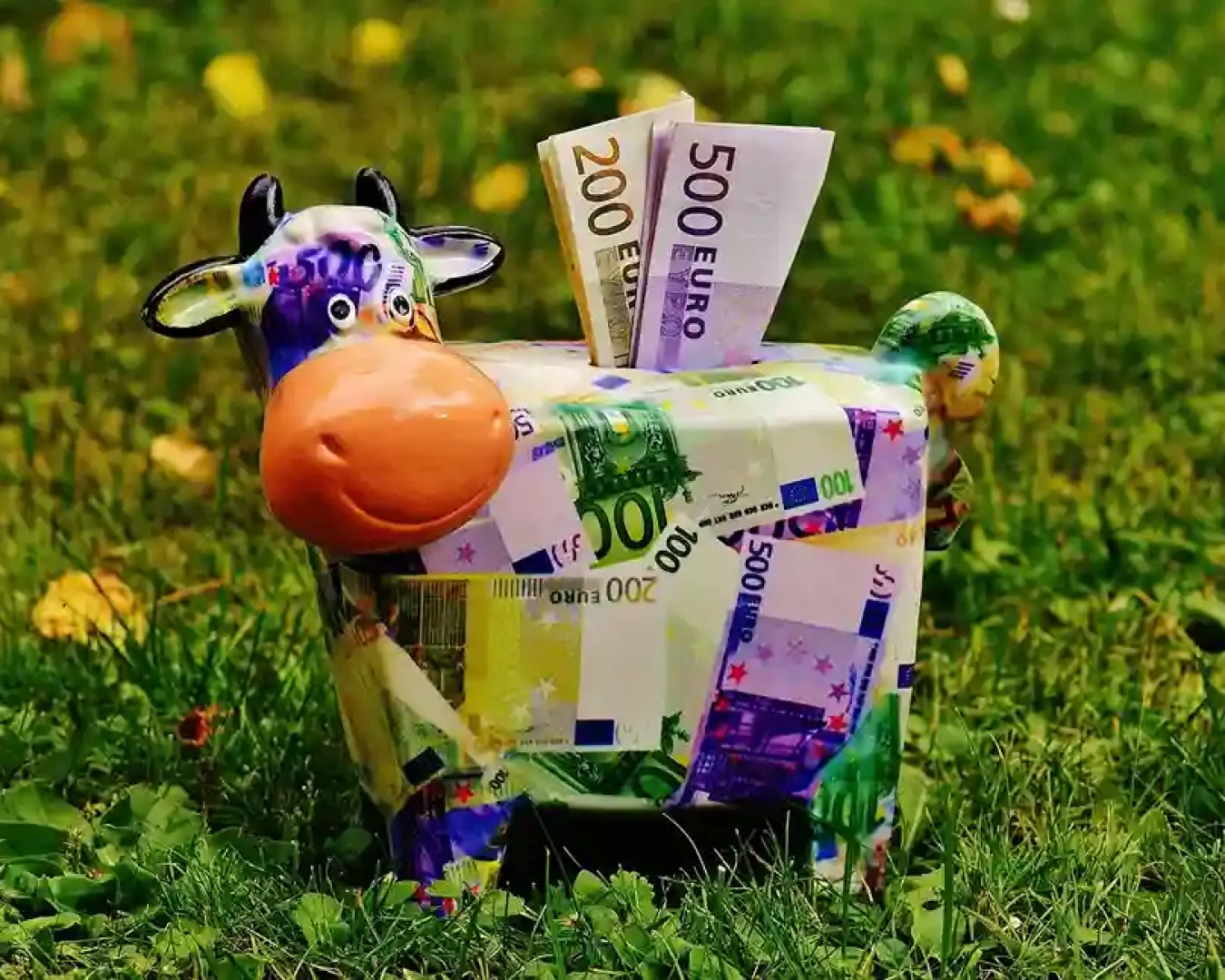 Ein farbenfrohes Sparschwein in Form einer Kuh, die mit Euro-Banknoten bedruckt ist, steht auf einer Wiese. Aus seinem Rücken ragen 500- und 200-Euro-Scheine heraus und symbolisieren die Wahl zwischen Tagesgeld und Festgeld beim Sparen.