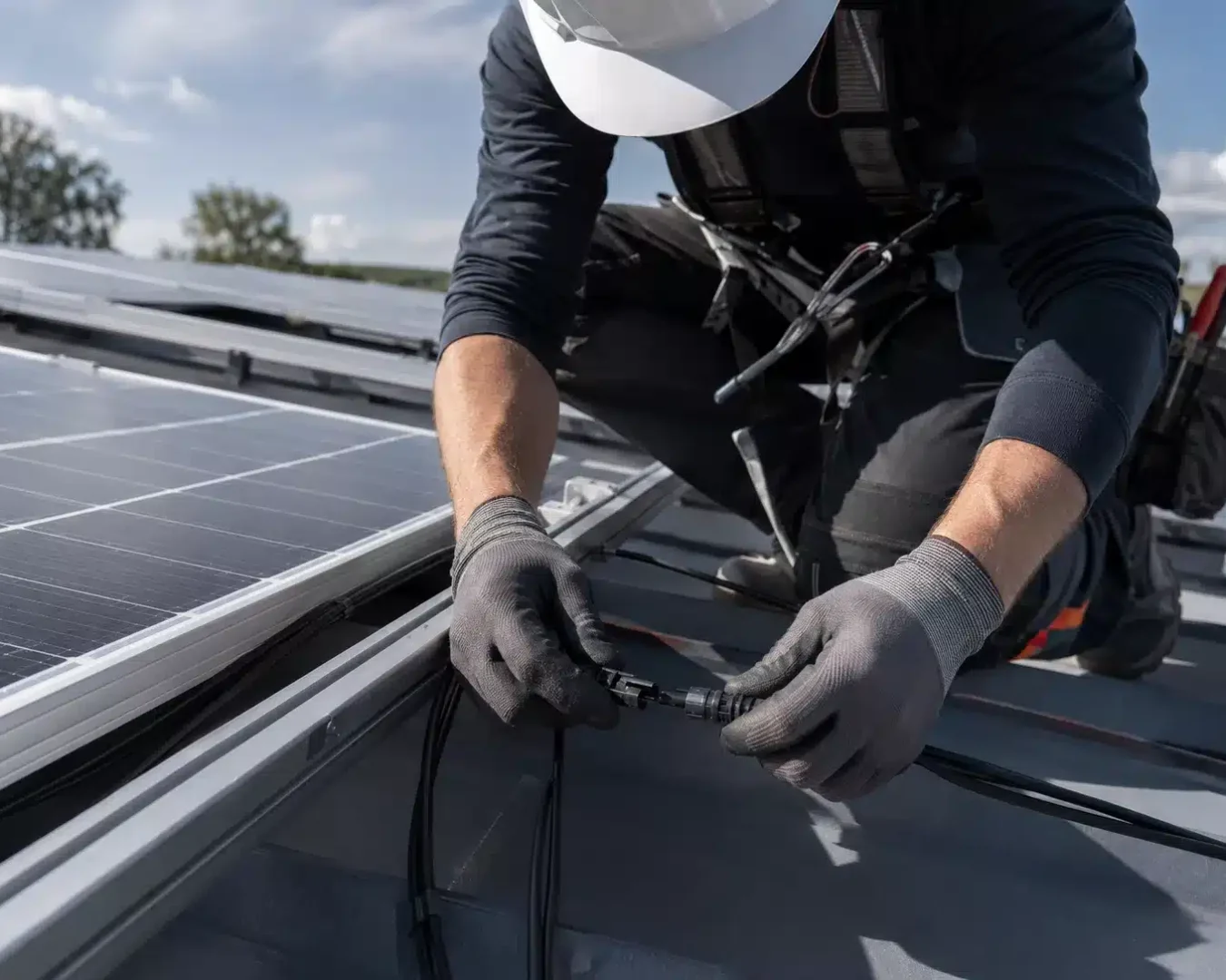 Ein Arbeiter, der Handschuhe und einen Helm trägt, schließt Kabel an ein Solarmodul auf einem Dach an und demonstriert dabei, wie man eine PV-Anlage fachgerecht anschließt – bei teilweise bewölktem Himmel.