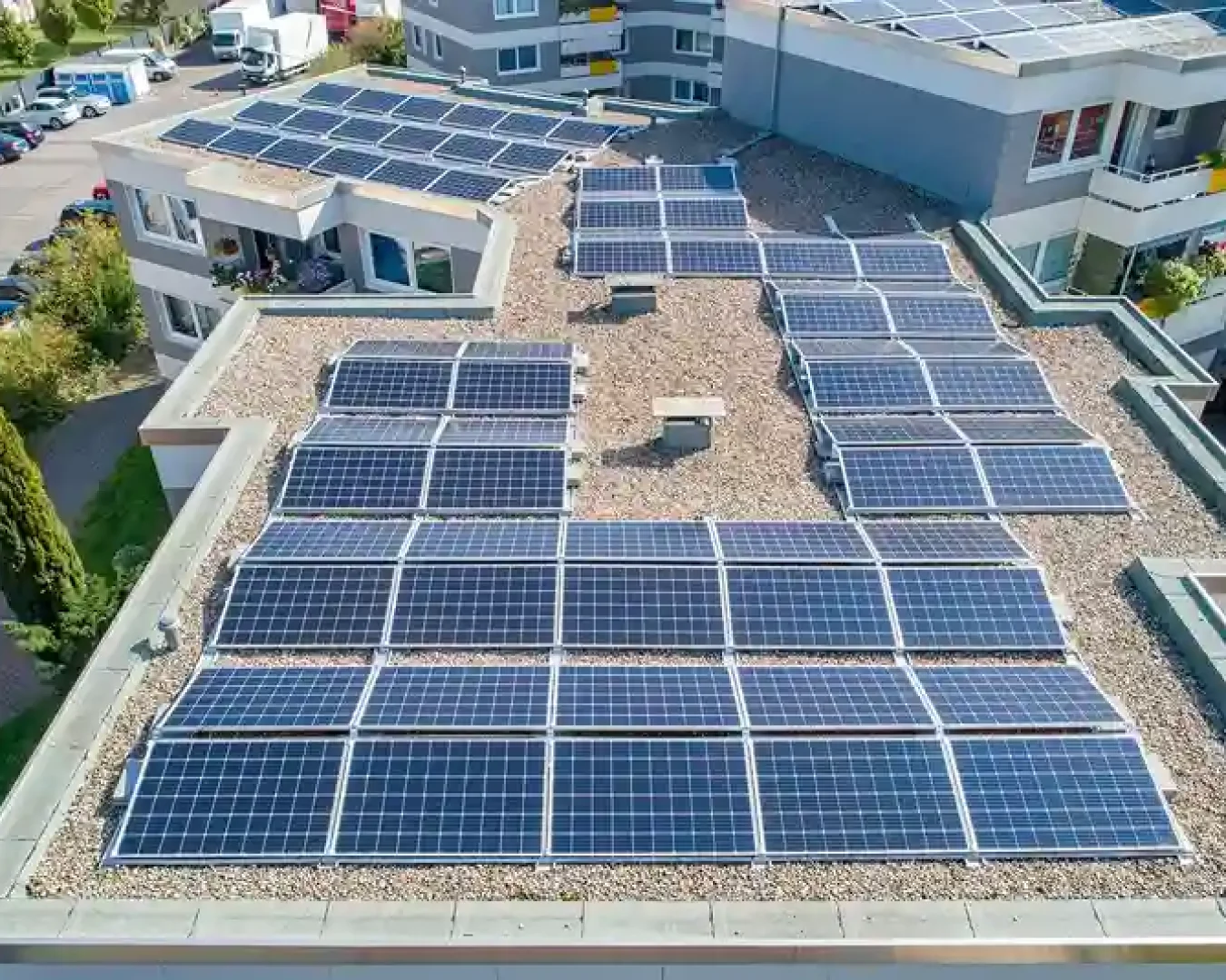 Luftaufnahme eines flachen, mit mehreren Solarmodulen bedeckten Daches eines modernen Gebäudes, das die Vorteile von Energiegenossenschaften gegenüber nachhaltigen Geldanlagen hervorhebt, umgeben von anderen Gebäuden, parkenden Autos und sichtbaren Grünflächen.