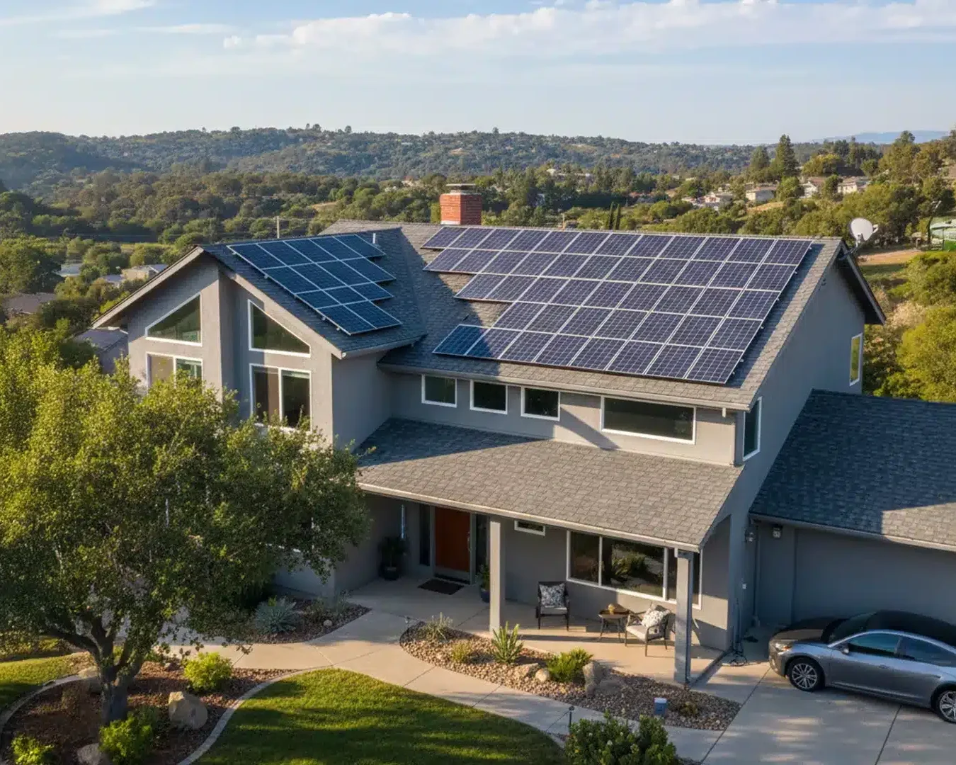 Ein modernes Haus mit Sonnenkollektoren auf dem Dach, umgeben von Bäumen und Grünflächen, mit Hügeln im Hintergrund und einem silbernen Auto, das an einem sonnigen Tag in der Einfahrt parkt.