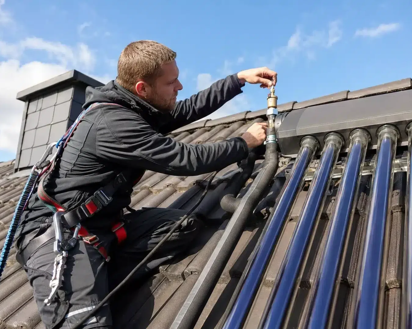Ein Arbeiter in Schutzkleidung führt unter blauem Himmel Wartungsarbeiten an einer Solaranlage auf einem Schrägdach durch und stellt dabei sorgfältig ein Ventil ein, das mit schwarzen Rohren und Glasröhren der Solarthermieanlage verbunden ist.