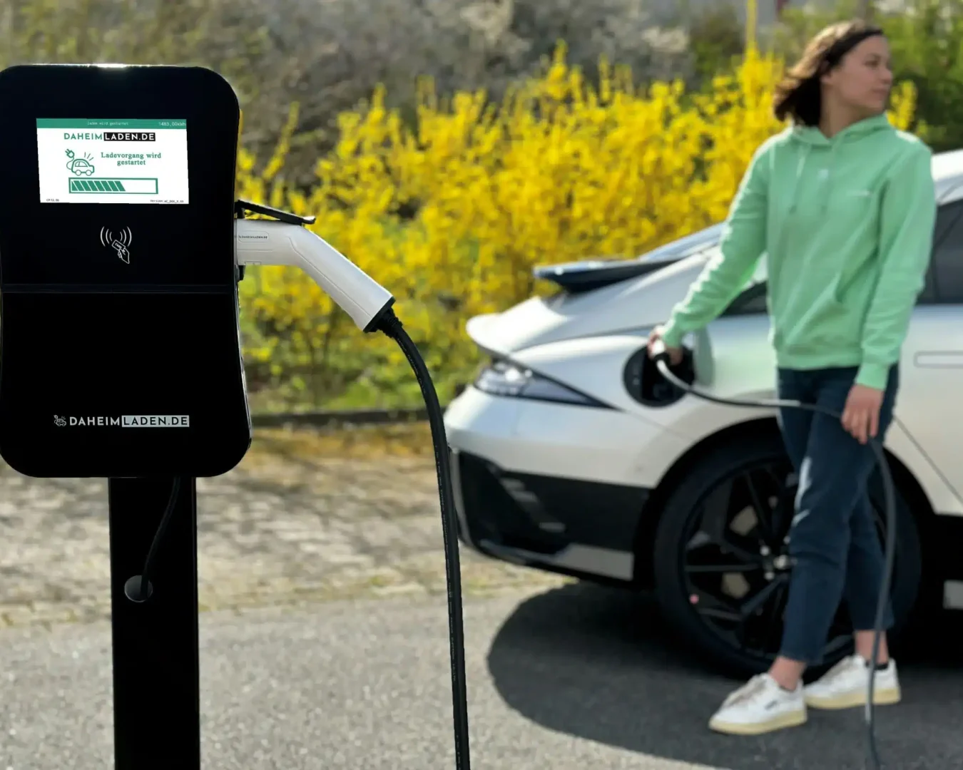Eine Wallbox steht vor einem weißen Auto und einer Frau. (Foto: Stephan Schwebe/Unsplash)